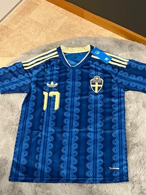 Blå Sverige fotbollströja Adidas - Säljer en blå fotbollströja från Adidas med Sveriges landslagsemblem och nummer 17 i gult. Tröjan har mönstrad design, gula detaljer och klassiska Adidas-ränder på axlarna. V-ringad krage och tillverkad i lätt, ventilerande material.
