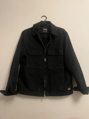 Svart overshirt från Zara - Svart overshirt från Zara med två stora bröstfickor och dubbel dragkedja framtill. Overshirten har klassisk krage och raka ärmar, tillverkad i ett kraftigt bomullstyg som ger en stilren look. Perfekt för lager-på-lager och streetstyle.