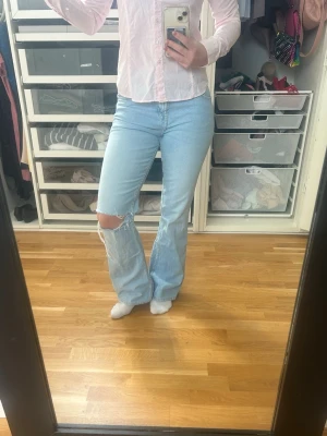 Ljusblå bootcut jeans med slitning - Säljer ett par ljusblå bootcut jeans med hål på vänster knä. Jeansen har klassiska fickor bak och fram, och är tillverkade i denim. Perfekt för dig som gillar en avslappnad och trendig look.