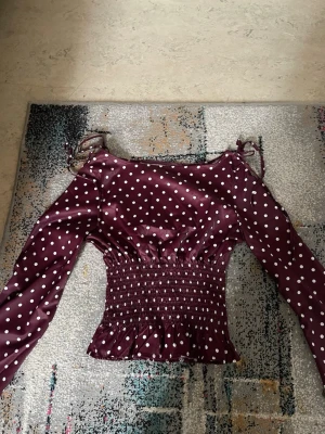Vinröd prickig topp med smock - Trendig vinröd topp med vita prickar. Toppen är HELT ny men den blev tyvärr för stor. Långärmad modell med smockad midja och volangkant nedtill. Knytband vid axlarna för extra detalj. Perfekt för dig som gillar feminina och unika plagg.
