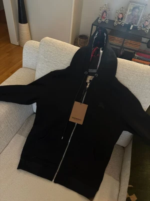 Svart hoodie från Burberry med dragkedja - Snygg svart hoodie från Burberry med klassiskt rutigt foder i huvan och dragkedja framtill. Märkets logga är diskret broderad på bröstet. Huvan har justerbara snören och materialet känns mjukt och bekvämt. Perfekt för dig som vill ha en exklusiv och stilren look. Aldrig använd med prislapp