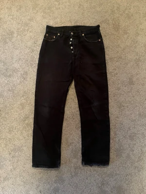 Svarta Levi's 501  - Säljer ett par klassiska svarta Levi's 501 jeans med raka ben och knappgylf. Jeansen har fem fickor, silverfärgade nitar och den ikoniska läderlappen bak i midjan. Storlek W33 L32. Hör av er vid fler funderingar✅