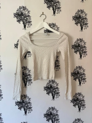 Ljus blommig långärmad topp - Ljus långärmad topp med små blommiga mönster och rundad, bred halsringning. Toppen är croppad och har en slim passform som framhäver siluetten. Perfekt för att skapa en söt och avslappnad look.