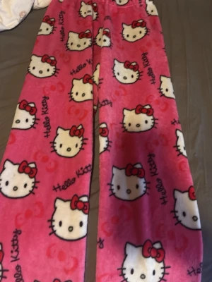 Hello Kitty rosa fleecebyxor - Mysiga loungewear-byxor i rosa fleece med Hello Kitty-motiv och röda rosetter. Byxorna har resår i midjan och är täckta av söta Hello Kitty-ansikten och texten 'Hello Kitty' tryckt i svart. Perfekta för chill dagar hemma.