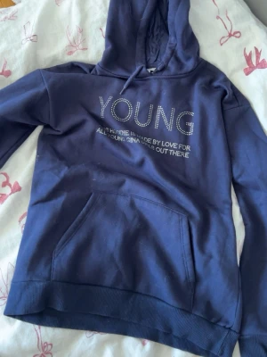 Mörkblå hoodie från Gina Tricot - Mörkblå hoodie med texten 'YOUNG' i silvriga nitar och tryck framtill. Klassisk känguruficka och ribbade muddar. Mjuk bomullskänsla och huva med dragsko. Perfekt för en chill och avslappnad stil.