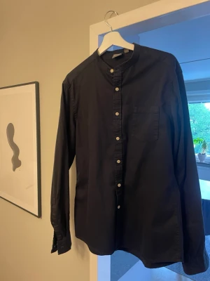 Svart henley-skjorta från ASOS - Svart henley-skjorta från ASOS med ståkrage och knappar framtill. Skjortan har lång ärm och bröstficka, tillverkad i mjuk bomull. Perfekt för en clean och stilren look. Storlek L