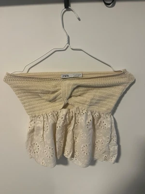 Beige offshoulder topp från Zara - Supersöt offshoulder topp från Zara i beige med broderade blommor och volang. Överdelen är smockad och ger en snygg passform, medan nederdelen är i ett lätt, broderat tyg. Perfekt för varma dagar och en trendig look.
