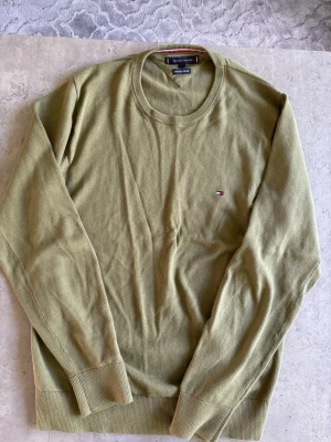 Olivgrön tröja från Tommy Hilfiger - Olivgrön rundhalsad tröja från Tommy Hilfiger med diskret logga broderad på bröstet. Tröjan har långa ärmar och ribbade muddar vid ärmslut och nederkant. Perfekt för en stilren och avslappnad look.
