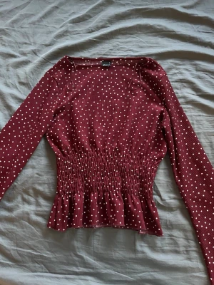 Prickig vinröd topp från Gina Tricot - Vinröd topp från Gina Tricot med vita prickar, långärmad och smockad midja som ger en snygg form. Toppen har volang nertill och är tillverkad i ett mjukt, stretchigt material. Perfekt för dig som gillar trendiga detaljer och bekväm passform.