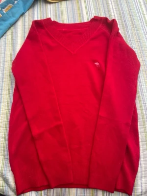 Röd stickad tröja Abercrombie & Fitch - Röd stickad tröja från Abercrombie & Fitch med V-ringning och ribbade muddar. Tröjan har en liten broderad älg-logga på bröstet och är tillverkad i mjukt material. Perfekt för dig som gillar klassisk och stilren design.