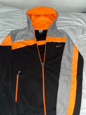 Nike vindjacka svart/orange/grå - Snygg vindjacka från Nike i svart, orange och grått. Jackan har huva, dragkedja framtill och en ficka med dragkedja. Nike-logga på bröstet och coola färgblock ger en sportig vibe. Perfekt för blåsiga dagar och streetstyle.