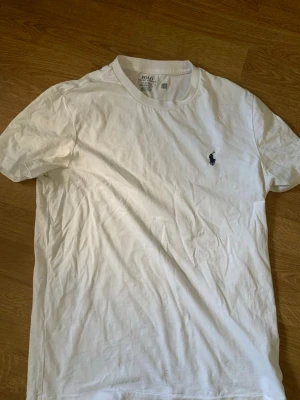  Ralph Lauren - Klassisk vit t-shirt från Polo Ralph Lauren med broderad marinblå logga på bröstet. T-shirten har rund halsringning och korta ärmar. Tillverkad i mjuk bomull för en skön känsla och stilren look. Har dock en fläck som kanske går bort om man tvättar men vet inte 