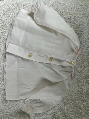 Vit blus med brodyr från Smock-Kläder - Söt vit babyklänning i str 70 cl, med puffärmar och broderad blomdetalj i blått och rosa framtill. Med  tre pärlemorknappar och rosa smock vid hals och ärmslut. Perfekt till babyns första sommar.