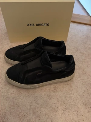 Svarta sneakers från Axel Arigato - Stilrena svarta sneakers från Axel Arigato med vit sula och diskret logga på sidan. Skorna har en låg silhuett och är tillverkade i mocka med elastisk panel över vristen istället för klassiska snören. Perfekta för en clean och modern look.