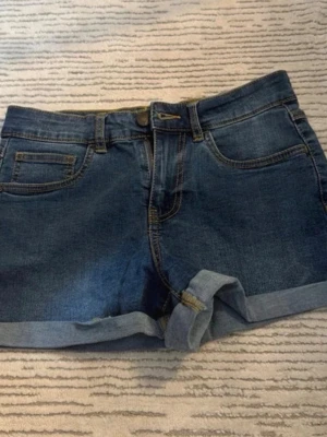 Blå jeansshorts med upprullad kant - Snygga blå jeansshorts med klassisk femficksdesign och upprullad kant nedtill. Perfekta för varma dagar och enkla att matcha med allt. Stängs med knapp och dragkedja framtill.
