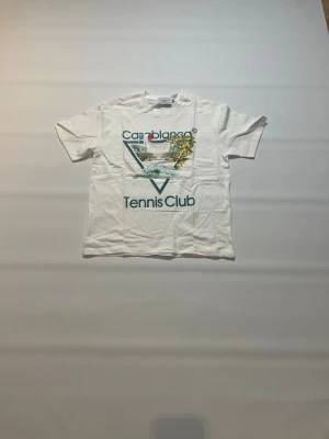 Vit Casablanca Tennis Club t-shirt storlek S - Vit t-shirt från Casablanca med tryck framtill där det står 'Tennis Club' och har en färgglad illustration. Klassisk passform med korta ärmar och rund hals. Perfekt för dig som gillar sportig och trendig stil till sommaren. Tröjan är helt ny och aldrig buren och har storlek S