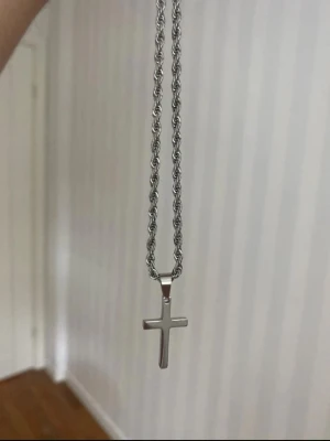 Silverfärgat ropechain halsband 60cm med kors - Stilrent halsband med ett kors som hänge. Kedjan har en tvinnad design som ger ett coolt och modernt intryck. Passar perfekt för dig som gillar enkla men snygga accessoarer. Materialet ser ut att vara metall.