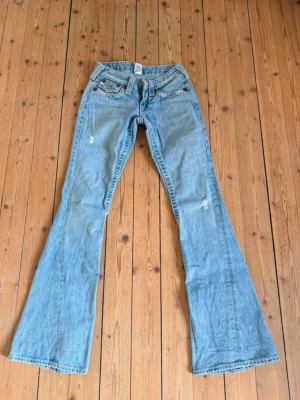 Ljusblå bootcut jeans från True Religion - Säljer ett par ljusblå bootcut jeans från True Religion med klassiska kontrastsömmar och slitningar på benen. Jeansen har låg midja, fem fickor och en snygg broderad detalj på bakfickan. Perfekta för dig som gillar retrovibbar och avslappnad stil. Går inte att få tag på längre, passar någon med Xxs eller Xs, se sista bild för att se att de är något slitna i benen och använt skick. ❤️
