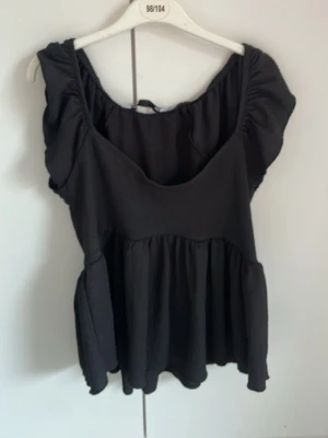 Svart blus med volangärmar - Supersöt svart blus med volangärmar och rundad halsringning. Toppen har en lös passform och är tillverkad i ett lätt, flowy material som ger en luftig känsla. Perfekt att styla med jeans eller kjol för en trendig look.