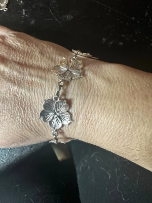 Silverfärgat armband med blommor - Snyggt armband i silverfärg med flera detaljerade blommor som länkas ihop. Blommorna har en tydlig struktur och armbandet har ett dekorativt lås med snirkliga detaljer. Perfekt för dig som gillar unika accessoarer med naturinspirerad design.