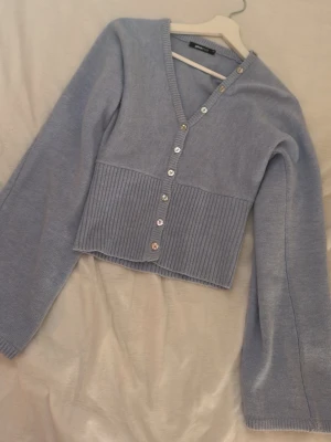 Ljusblå kofta från Gina Tricot - Supersöt ljusblå kofta från Gina Tricot med v-ringning och glansiga knappar framtill. Modellen är croppad med bred ribbstickad nederdel och långa ärmar. Perfekt att styla med jeans eller kjol för en trendig look.