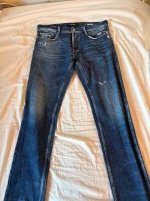 Blå jeans från Replay, modell Anbass - Säljer ett par snygga blå jeans från Replay, modell Anbass. Jeansen har en slim fit-passform, klassisk femficksdesign och slitna detaljer framtill för en cool look. Tillverkade i stretchigt denim för extra komfort.