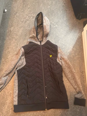 Grå fleece-tröja från Lyle & Scott - Mysig grå fleece-tröja från Lyle & Scott i storlek M. Tröjan har en melerad utsida och mjuk fleece på insidan, perfekt för kyliga dagar. Enkel och stilren design som passar till många outfits. Har bara fått en litet hål som du ser