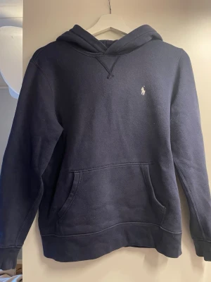Mörkblå hoodie från Polo Ralph Lauren - Snygg mörkblå hoodie från Polo Ralph Lauren med klassisk huva och känguruficka. Liten vit broderad logga på bröstet. Tillverkad i mjukt bomullsmaterial som känns skönt mot huden. Perfekt för en avslappnad och stilren look. Väldigt bra skick! Stoleken är L men är från kids brand store så skulle säga att stoleknen är xs! 