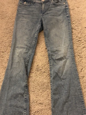Ljusblå bootcut jeans med detaljer - Säljer ett par ljusblå bootcut jeans med snygga broderade bakfickor och silvriga  knappar. Jeansen har klassisk femficksdesign och är tillverkade i denim med lätt tvättad look. Perfekta för en avslappnad och trendig stil.