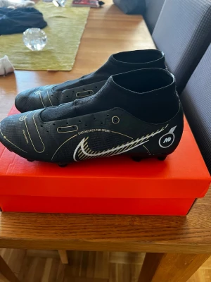 Svarta Nike Mercurial fotbollsskor - Snygga svarta Nike Mercurial fotbollsskor med vita och guldiga detaljer. Skorna har högre strumpliknande skaft för extra stöd och är designade för fart på planen. Syntetiskt material med klassisk Nike Swoosh på sidan och mönstrad yttersula för bra grepp.