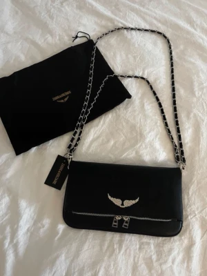 Svart Zadig & Voltaire axelväska - Snygg svart axelväska från Zadig & Voltaire i läder med silverfärgad kedjerem och ikonisk vinglogga framtill. Väskan har dragkedjedetalj och stilren design, perfekt för att lyfta din outfit. Lånad Bild! 
