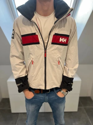 Helly Hansen vit och röd vindjacka - Hej säljer nu en snygg vit vindjacka från Helly Hansen med röda och svarta detaljer. Jackan har dragkedja, huva och två fickor framtill. Stora HH-loggor på bröstet och huvan. Perfekt för blåsiga dagar och streetwear-vibe. Lagning på höger ärm, syns på bild fyra.  Super att ha nu inför regn, Storlek M men något mindre i storlek så mer åt S hållet och pris 650kr men kan gå ner vid snabb affär💙💯📦 