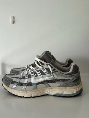Nike P-6000 grå sneakers - Nike P-6000 sneakers i grått och silver med vita detaljer. Ovandel i mesh och syntetmaterial, med tydliga paneler och reflekterande inslag. Klassisk Nike-logga på sidan och tungan. Skön och sportig sula i beige och svart. Perfekta för en streetwear-look. OBS LÅDA tillkommer även KVITTO VID FÖRFRÅGAN 