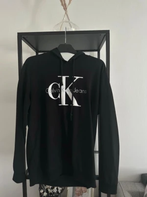 Svart hoodie från Calvin Klein Jeans - Snygg svart hoodie från Calvin Klein Jeans med stort vitt CK-tryck och logga på bröstet. Klassisk modell med huva och dragsnören, samt långa ärmar. Tillverkad i mjukt material som passar perfekt till en avslappnad stil. Tyvärr finns inte storleks lappen kvar. På CKs hemsida visar det att den ska vara i storlek M/L. Måtten längst ner på hoodien är 105cm. På mig slutar den i mitten av rumpan, är 170cm lång 💗