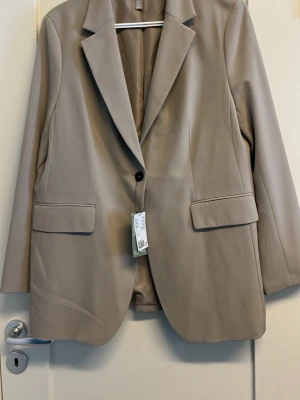 Beige fodrad kavaj från H&M - Ny Stilren beige kavaj med lätt stretch från H&M med klassisk krage, en knapp framtill och två fickor med lock. Långa ärmar med knappar vid ärmslut. Perfekt för en trendig och avslappnad look.       Yttertyg: 96 % polyester 4% elastan  fodertyg: 100%polyester Aldrig använd  Nypris: 599:-