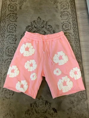 Rosa shorts med vita blommor - Säljer ett par rosa shorts med vita blommönster och snörning i midjan. De har avslappnad passform, elastisk midja och är tillverkade i mjukt bomullstyg. Perfekta för sommarens alla äventyr!