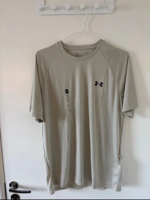 Beige träningströja från Under Armour - Oanvänd med prislapp, nypris: 349kr Säljer en beige kortärmad träningströja från Under Armour i storlek M. Tröjan har en lös passform, rund halsringning och är tillverkad i ett lätt och snabbtorkande funktionsmaterial. Perfekt för gymmet eller löprundan.