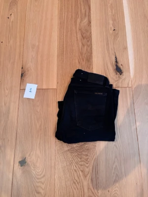 Svarta slim fit jeans från Nudie - Säljer ett par svarta slim fit jeans från Nudie Jeans. Jeansen har klassisk femficksdesign, svarta sömmar och en diskret logga på bakfickan. Materialet är stretchigt och bekvämt, perfekt för en snygg och stilren look.