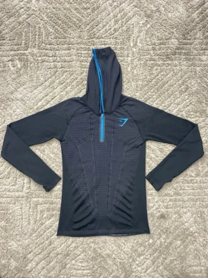 Gymshark Onyx V1-hoodie - Sportig svart hoodie från Gymshark med lång ärm och halv dragkedja i blått framtill. Tröjan har ett mönstrat parti på bröst och rygg samt Gymshark-logga på vänster bröst. Tillverkad i ett stretchigt funktionsmaterial som andas. 
