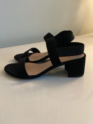 Svarta sandaler med bred klack H&M - Stilrena svarta klackar från H&M med bred klack och öppen tå. Klackarna har en bred rem över foten och elastiskt band runt hälen för extra komfort. Ovandelen är i mockaimitation och innersulan är beige. Perfekta till sommaren!