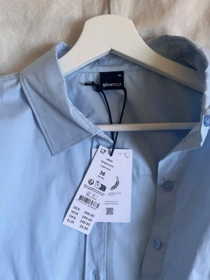 Ljusblå skjorta från Gina Tricot - Kortärmad ljusblå skjorta från Gina Tricot med klassisk krage och knappar framtill. Skjortan har ett knytband i midjan för en snygg passform och är gjord i mjuk bomull. Perfekt för en fräsch och stilren look.
