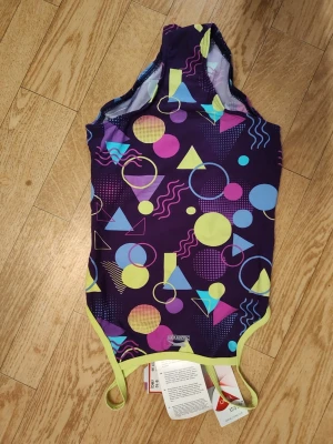 Färgglad baddräkt från Slazenger stl 134-140 / 9-10 - Säljer en cool baddräkt från Slazenger med lila bas och färgglada geometriska mönster i gult, rosa, blått och turkost. Baddräkten har kontrasterande ljusgula detaljer och axelband. Perfekt för dig som gillar att sticka ut på stranden eller i simhallen. Barn stl 9-10/134-140. Helt ny oanvänd! 