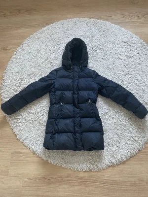 Svart lång dunjacka från Gant - Snygg svart lång dunjacka från Gant med huva och dragkedja framtill. Jackan har två fickor med dragkedja och quiltad design som ger en klassisk vinterlook. Perfekt för kalla dagar då den är väldigt varm! Storlek S🥰kostar ca 7000 nypris