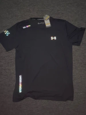 Svart träningströja från Under Armour - Snygg svart t-shirt från Under Armour med regnbågsfärgade loggor och textdetaljer. Tillverkad i ett lätt och snabbtorkande funktionsmaterial som passar perfekt till träning. Rund halsringning och korta ärmar för maximal rörelsefrihet.