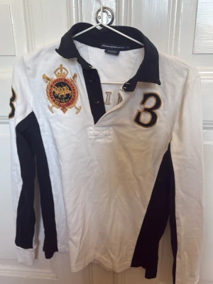 Vit och svart henley från Ralph Lauren - Snygg långärmad rugby från Ralph Lauren i vitt och svart med broderad logga och siffran 3 på bröstet och ryggen. Skjortan har klassisk krage, knappar framtill och detaljer i guld och svart. Perfekt för dig som gillar  stilren look.