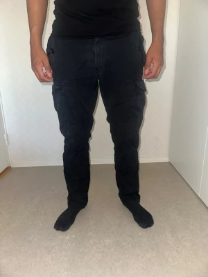 Ralph lauren Svarta cargobyxor med fickor - Säljer ett par svarta cargobyxor med flera praktiska fickor och knappdetaljer. Byxorna har en rak passform och är tillverkade i ett slitstarkt bomullstyg. Perfekta för en avslappnad och cool stil.   Person i bild är 183cm lång