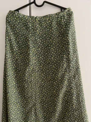 Grön blommig långkjol (below knee A line skirt) - Snygg långkjol i mörkgrönt med småblommigt mönster i ljusare toner. Kjolen har en lätt utställd form och är perfekt för dig som gillar en avslappnad men ändå trendig stil. Materialet känns mjukt och följsamt mot huden.