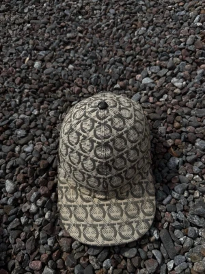 Ferragamo keps - Snygg beige keps med svart grafiskt mönster över hela ytan. Klassisk böjd skärm och knapp på toppen. Justerbar rem baktill för perfekt passform. Perfekt accessoar för en trendig streetwear-look.