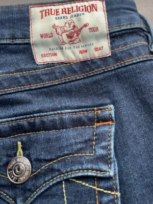 True Religion blå jeans med fickdetaljer - Säljer ett par klassiska blå jeans från True Religion med snygga kontrastsömmar och ikoniska fickdetaljer bak. Jeansen har bootcut-modell och coola knappar med logga. Perfekta för dig som gillar streetstyle och vill sticka ut.