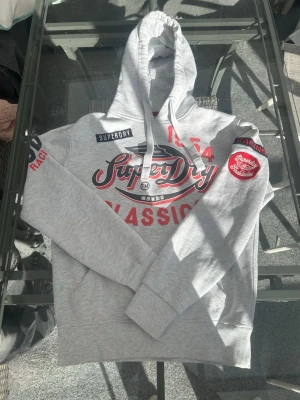 Superdry Racing Hoodie – Y2K Streetwear Grey/Red - Säljer en riktigt snygg Superdry hoodie i ljusgrå färg med racing-inspired graphic print och klassiska detaljer som ger en clean vintage och y2k streetwear look. Hoodien har mjuk fleecefodrad insida vilket gör den perfekt för både casual wear, vardag och chill sommarkvällar.  Passar perfekt med jeans, cargos eller shorts för en modern streetstyle fit. Superdrys japansk-inspirerade design och old school racing aesthetic gör att den sticker ut direkt.  Storlek S och i mycket bra skick.